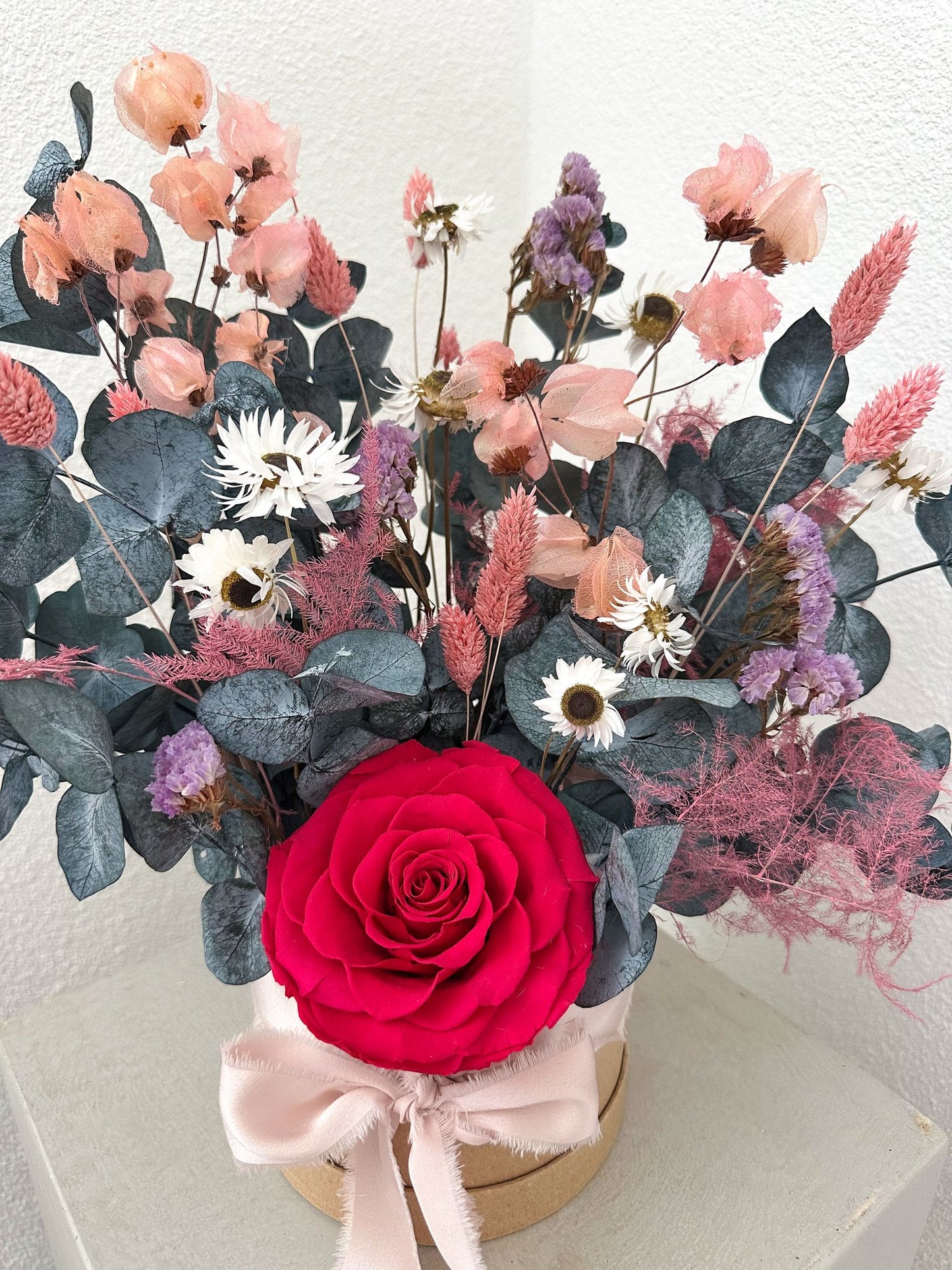 Arrangement mit echter Rose & Trockenblumen auf hellem Sockel – harmonisch komponiert in Rosé, Bordeaux und Eukalyptusgrün.