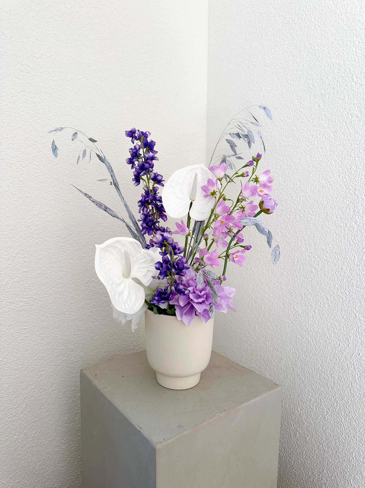 Kunstblumenarrangement „Mary“ in weisser Vase mit Pfingstrosen, Delphinium, Campanula und Chasmanthium auf hellem Sockel.