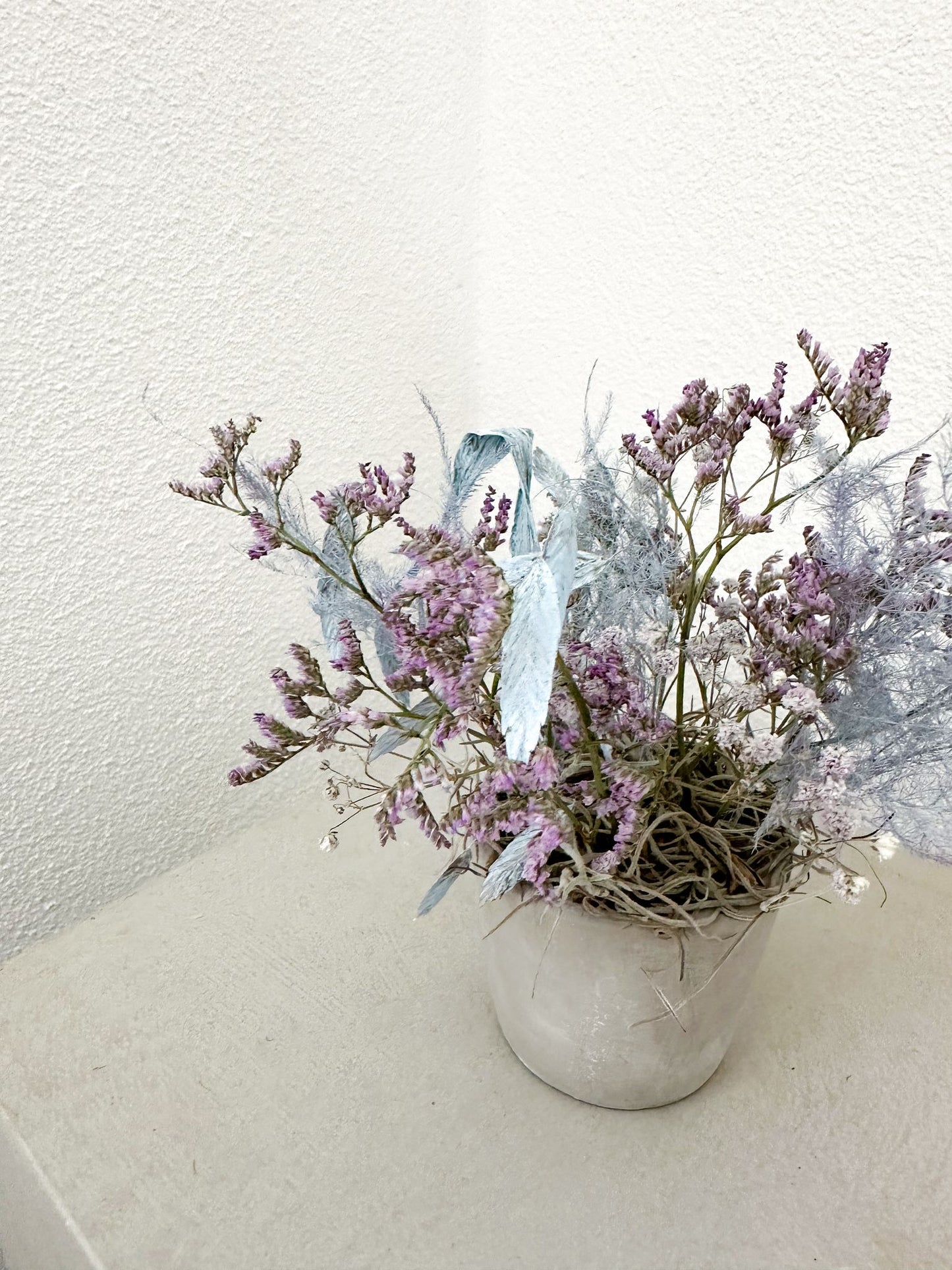 Blumengesteck Sina auf Betonsockel in ruhiger Ecke – sanftes Farbschema aus Lavendel, Nebelblau und Grau.