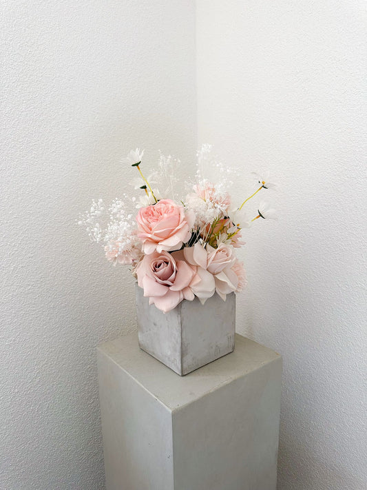 Romantisches Kunstblumenarrangement mit rosafarbenen Rosen, Margeriten und Schleierkraut in Betonwürfel – zart & verspielt inszeniert.