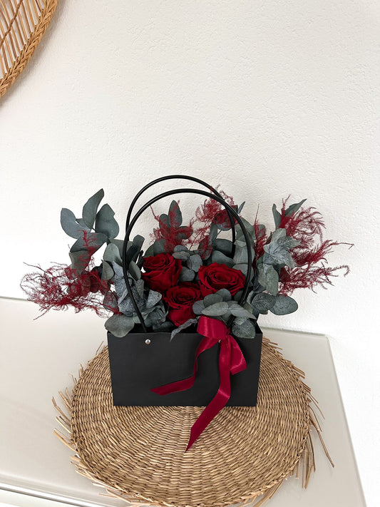 Elegante Blumentasche mit stabilisierten roten Rosen, grauem Eukalyptus und filigranen Farnen, dekoriert mit rotem Satinband.