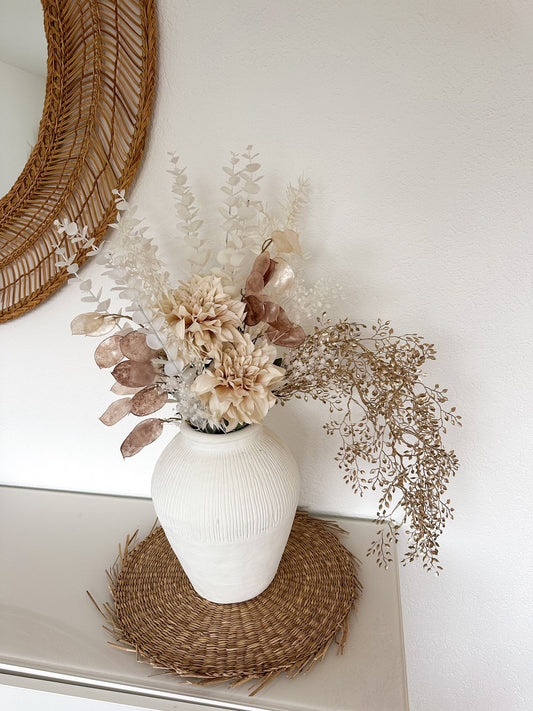 Carina Bouquet in einer weissen Vase, präsentiert auf einer geflochtenen Unterlage vor einem dekorativen Wandspiegel.