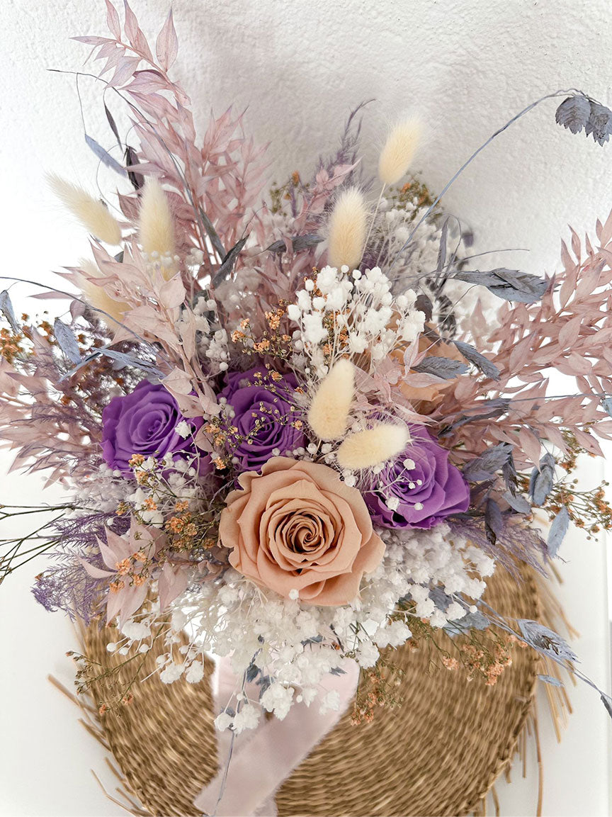 Zauberhafter Anblick des Mama Bouquet, kunstvoll arrangiert mit lila und cremefarbenen Trockenblumen auf natürlicher Rattanunterlage.