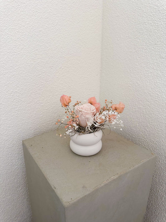 Zartes Mini-Gesteck „Simona“ aus Trockenblumen mit stabilisierter Rose, Bougainvillea, Ruscus und Schleierkraut auf hellem Podest.